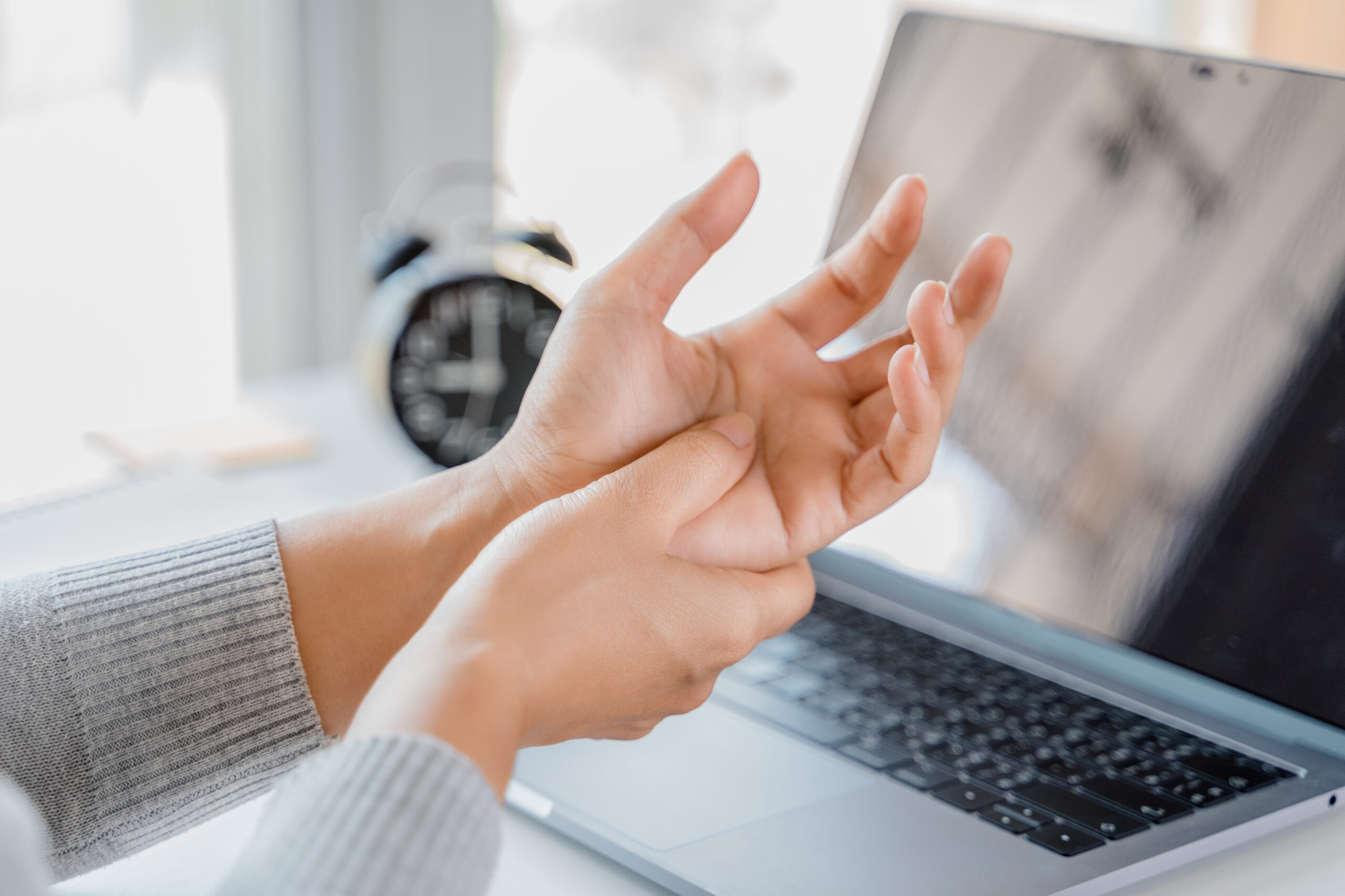Woman holding her wrist and hand in pain while using a laptop, showing signs of hand pain and repetitive strain injury from prolonged computer use.