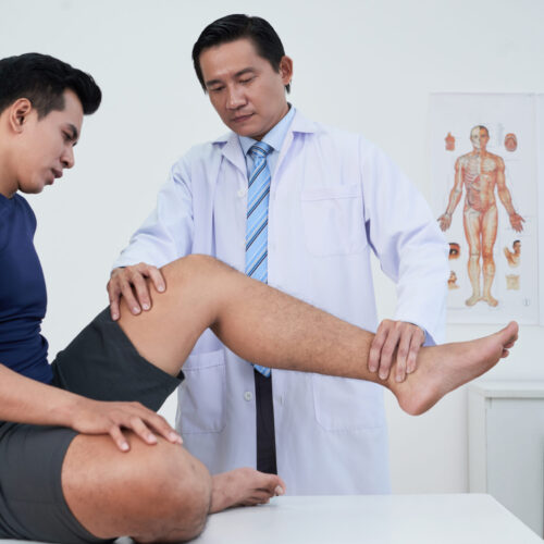 A physiatrist performs a physical examination on a male athlete’s knee in a clinical setting, checking for joint stability and signs of injury.