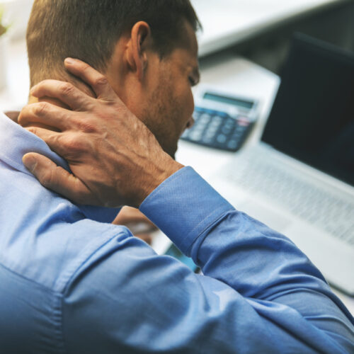 Man sitting at a desk in front of a computer, holding his lower back in pain—depicting discomfort from long hours of sitting or poor posture.
