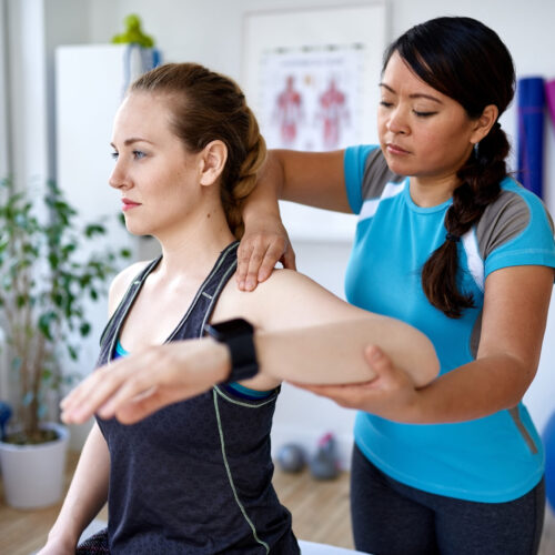 Physical therapist guides a patient in a bright medical office through a hands-on mobility exercise, focusing on shoulder and upper body flexibility.