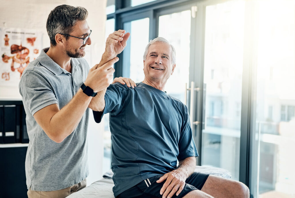 With a smile, a senior man performs arm-exercises under the guidance of a physical therapist in a rehabilitation clinic, focusing on improving strength and mobility.