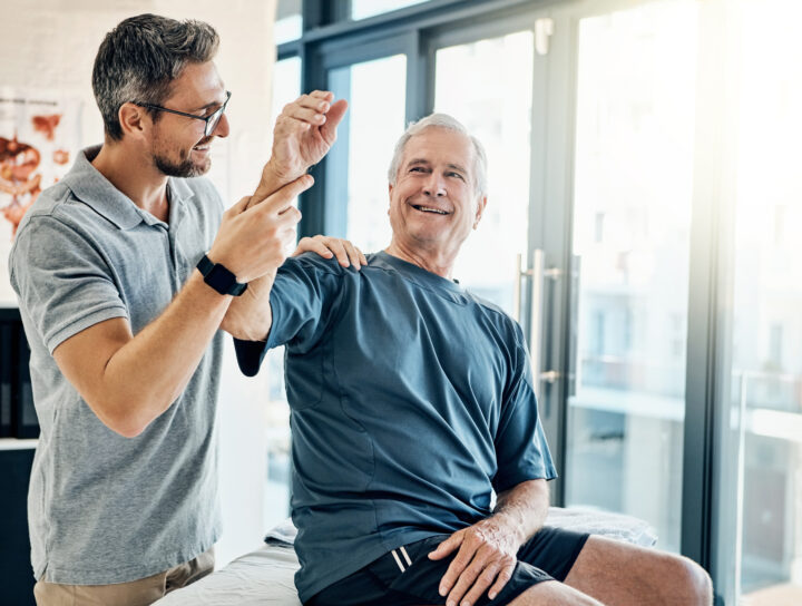 With a smile, a senior man performs arm-exercises under the guidance of a physical therapist in a rehabilitation clinic, focusing on improving strength and mobility.