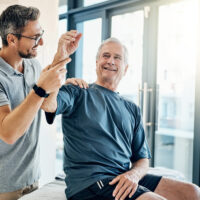 With a smile, a senior man performs arm-exercises under the guidance of a physical therapist in a rehabilitation clinic, focusing on improving strength and mobility.