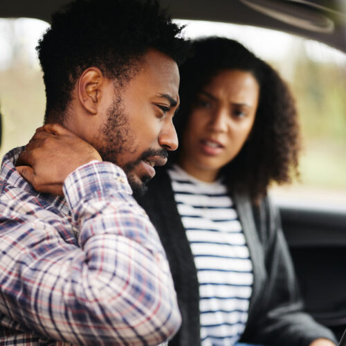 Man in car holding his neck after a traffic accident, showing delayed neck pain or whiplash injury while passenger looks on with concern.