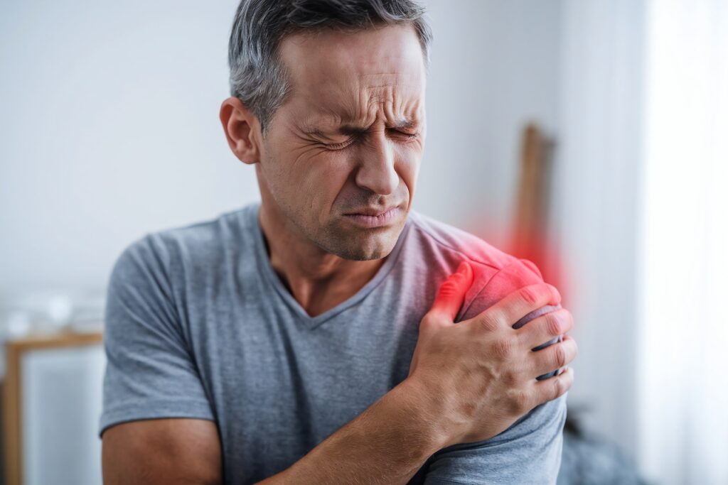 Middle-aged man sitting at home holding his shoulder, showing signs of shoulder blade pain.