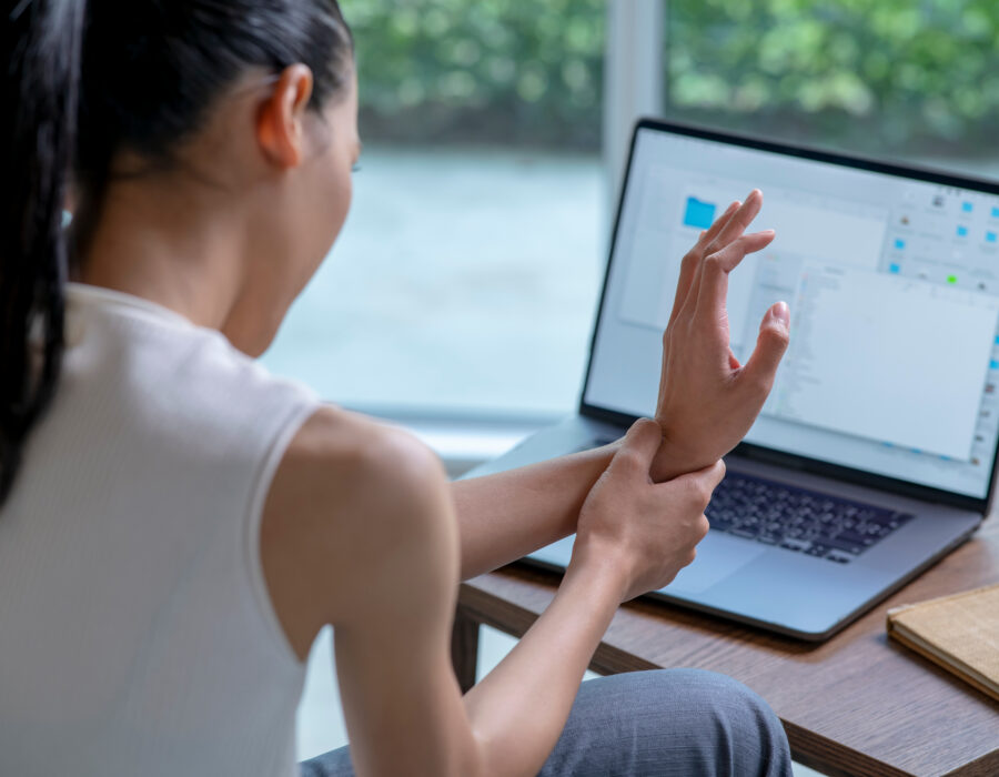 Woman working on laptop at home holding wrist in pain, showing possible carpal tunnel syndrome symptoms.
