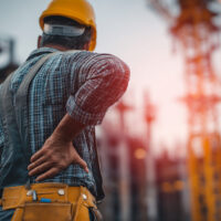 Construction worker holding his lower back in pain at a job site, representing back pain after a construction injury.