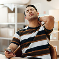 Man holding the back of his neck while looking at his phone at a desk, illustrating tech neck and neck pain from prolonged device use.