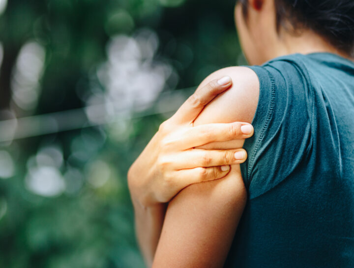 Person outdoors holding shoulder and upper arm, illustrating dull vs sharp pain in the shoulder area possibly related to muscle strain or overuse.