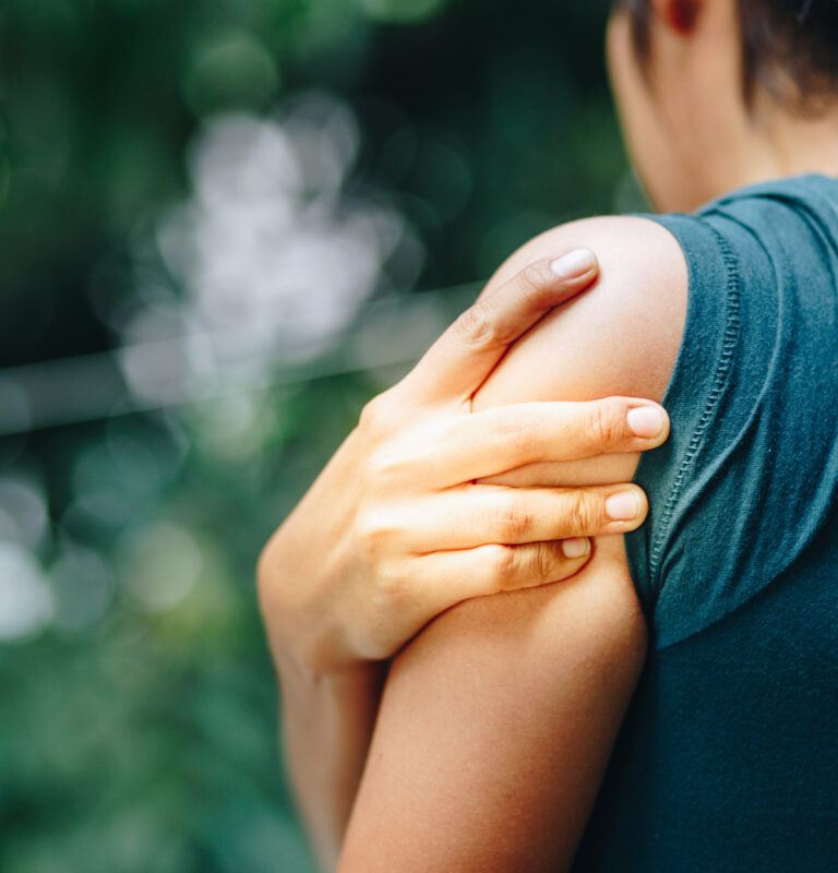 Person outdoors holding shoulder and upper arm, illustrating dull vs sharp pain in the shoulder area possibly related to muscle strain or overuse.