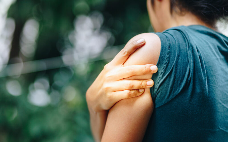 Person outdoors holding shoulder and upper arm, illustrating dull vs sharp pain in the shoulder area possibly related to muscle strain or overuse.