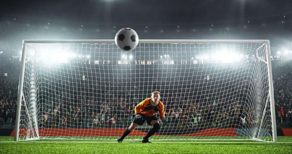 Soccer goalkeeper preparing to defend a shot on goal during a match, illustrating the unique physical demands and injury risks associated with the goalkeeper position.